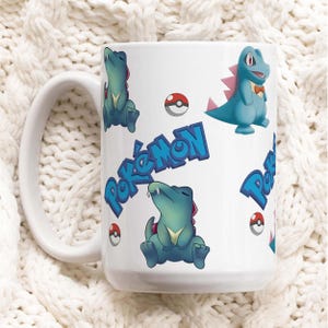 Può includere: Tazza in ceramica bianca con un motivo ripetuto di coccodrilli dei cartoni animati blu e verdi, grafiche di Pokeball rosse e bianche e la parola "Pokémon" in blu. La tazza ha un manico grande e curvo.