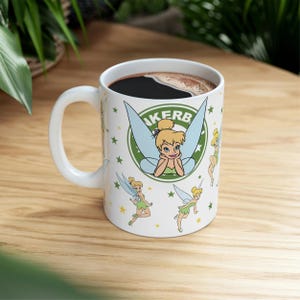 Op de afbeelding: Witte keramische koffiemok met Tinkerbell-illustraties en het woord "KERB" in een groene cirkel. De mok is gevuld met zwarte koffie en staat op een houten oppervlak.