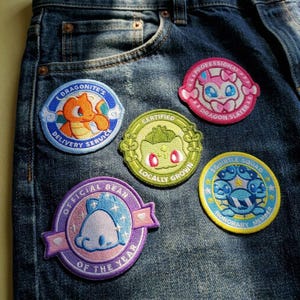 Puede incluir: Cinco parches bordados circulares con personajes de dibujos animados y texto sobre tela vaquera azul oscuro. Los parches incluyen Dragonite's Delivery Service, un Dragon Slayer rosa, un Bulbasaur verde, un Official Bean morado y un Squirtle Squad.