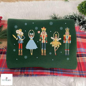 May include: A dark green sweatshirt featuring embroidered Nutcracker-themed characters including a mouse king, a ballerina in a blue dress, a nutcracker soldier, a fairy, and a traditional soldier, all surrounded by small white snowflake accents.