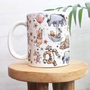 Op de afbeelding: Witte keramische mok met illustraties van Winnie de Poeh en zijn vrienden. Het ontwerp bevat personages als Poeh, Knorretje, Tijgertje en Iejoor, samen met bloemen en een roze ballon. De mok heeft een wit handvat.