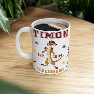 Può includere: Tazza in ceramica bianca con il personaggio Timon de Il Re Leone. La tazza presenta la scritta "TIMON" in lettere marroni, il testo "EST. 1994" e le parole "THE LION KING". La tazza è riempita di liquido scuro.