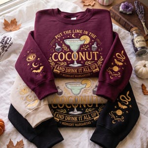 Könnte beinhalten: Ein Stapel aus drei Rundhals-Sweatshirts in Kastanienbraun, Creme und Schwarz. Jedes zeigt eine Grafik eines Margaritaglases mit dem Text "PUT THE LIME IN THE COCONUT AND DRINK IT ALL UP" und "MIDNIGHT MARGARITAS" sowie Ärmeldesigns mit Augen, Monden, Fledermäusen, Schlangen und Planeten.