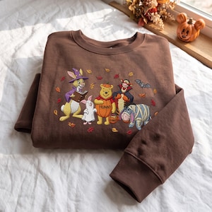May include: A brown crewneck sweatshirt featuring an embroidered graphic of Winnie the Pooh characters dressed in Halloween costumes, including a rabbit in a wizard hat, a piglet as a ghost, Pooh as a pumpkin, Tigger as a vampire, and Eeyore as a mummy.