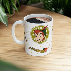 Könnte beinhalten: Weiße Keramiktasse mit Peter Pan-Illustrationen. Die Tasse hat einen großen Henkel und ist mit dunkler Flüssigkeit gefüllt. Das Design enthält den Text "PETER" in einem grünen Kreis mit einem Cartoon-Bild von Peter Pan und goldenen Sternen.