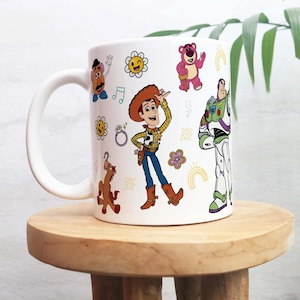Può includere: Tazza in ceramica bianca con personaggi di cartoni animati colorati, tra cui Woody e Buzz Lightyear. La tazza ha un manico bianco ed è decorata con fiori e arcobaleni. La tazza è appoggiata su una superficie di legno. Altezza della tazza: circa 9,5 cm.