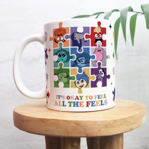 Könnte beinhalten: Weißer Keramikbecher mit Henkel, verziert mit Cartoonfiguren in Puzzleteilformen. Der Becher trägt den Text "IT'S OKAY TO FEEL ALL THE FEELS". Der Becher steht auf einer Holzoberfläche.
