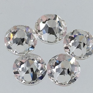 May include: Six clear round glass rhinestones with a faceted design. The rhinestones are arranged in a circle on a white surface.