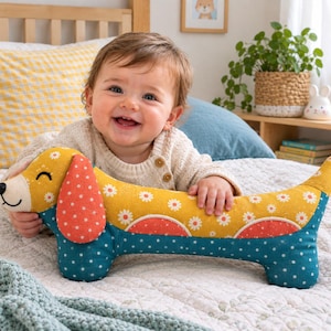 Puede incluir: Un juguete de peluche con forma de perro salchicha que presenta un lomo amarillo con un patrón de margaritas blancas, un cuerpo inferior verde azulado con lunares blancos y orejas color coral con lunares blancos sobre tela de color crema.