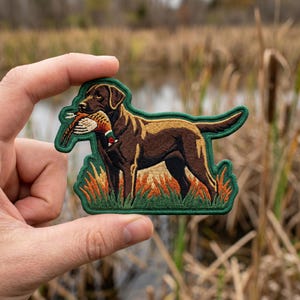 May include: An embroidered patch featuring a brown Labrador retriever standing in tall grass, holding a pheasant in its mouth. The patch has a dark green border and depicts the dog with detailed fur shading and a colorful bird in its jaws.