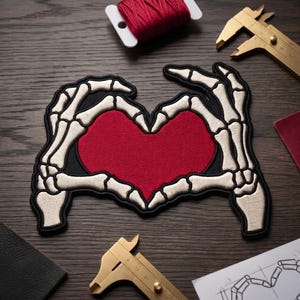 May include: Embroidered patch featuring a red heart shape framed by skeleton hands. The patch has a black border and off-white bone details. The design is on a dark wooden surface, with red thread and calipers in the background.