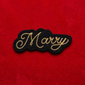 May include: A black felt patch with the word "Marry" embroidered in gold thread. The patch has a cloud-like shape and is set against a red background.