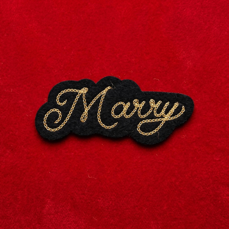 May include: A black felt patch with the word "Marry" embroidered in gold thread. The patch has a cloud-like shape and is set against a red background.