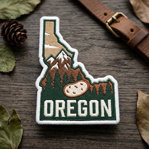 May include: Embroidered patch in the shape of the state of Oregon, featuring a mountain range, trees, and a potato. The patch is bordered in white with the word "OREGON" in white block letters.