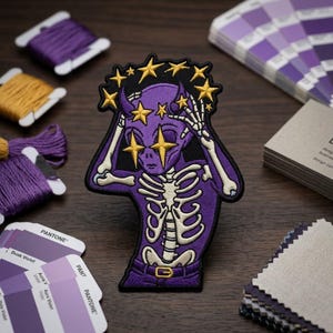 May include: Embroidered patch featuring a purple alien with a skeleton design, gold star accents, and a star crown. The patch is surrounded by purple thread spools, color swatches, and fabric samples on a dark wood surface.