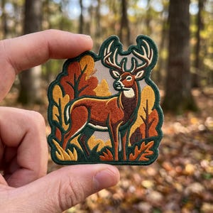 May include: An embroidered patch featuring a brown deer with large antlers standing in a forest setting with stylized orange, yellow, and dark green trees and foliage. The patch has a dark green border and a light-colored background texture.