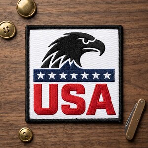 May include: Embroidered patch featuring a black eagle head above a blue stripe with white stars and the red letters "USA" on a white background. The patch has a black border and is on a wooden surface.