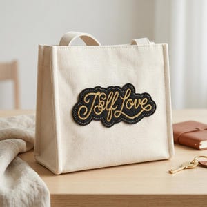 May include: A cream-colored canvas tote bag with a black patch that reads "Self Love" in gold script. The bag has a rectangular shape with a handle and is sitting on a wooden surface. A notebook and keys are also visible.