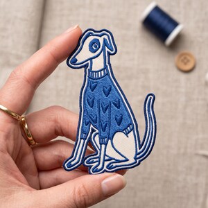 May include: An embroidered patch featuring a white dog with a blue nose and eye patch, wearing a blue sweater decorated with a repeating heart pattern. The dog is depicted in a seated position with its tail curved upward, outlined in dark blue thread.