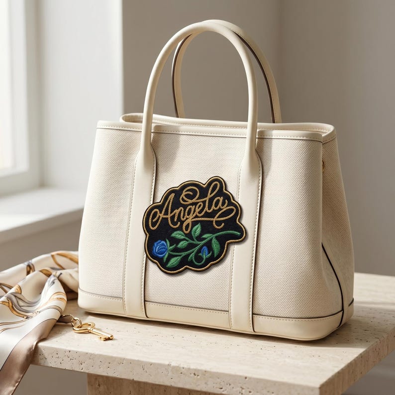 Pu&ograve; includere: Borsa tote color crema con una toppa ricamata nera e oro con la scritta "Angela" e un motivo floreale. La borsa ha due manici e un bordo color crema. Una sciarpa e una chiave sono sul tavolo.