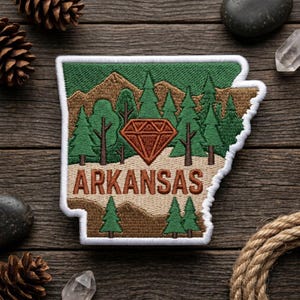 May include: Embroidered patch in the shape of Arkansas, featuring a diamond, trees, and mountains. The word "ARKANSAS" is stitched in brown. The patch has a white border and is set against a wooden background.