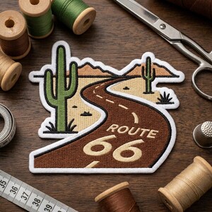 May include: An embroidered patch featuring a desert landscape with two green cacti, tan mountains, and a brown road winding through the scene. The text ROUTE 66 is stitched in tan thread on the road surface, surrounded by a white border.