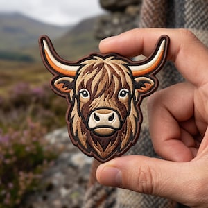 May include: An embroidered patch featuring the head of a Highland cow with long, curved horns in shades of cream and orange. The cow has shaggy brown fur, dark eyes, and a light-colored muzzle, all outlined with a dark brown border.