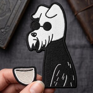 May include: An embroidered patch featuring a stylized black and white dog with a beard and round black sunglasses. The dog is depicted in profile, facing left, with a black body and white head. A small white coffee cup is positioned near the dog.