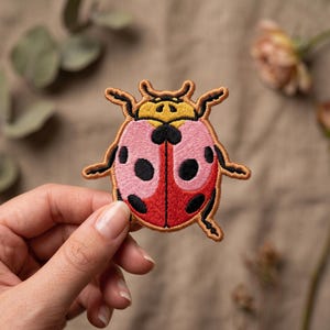 May include: An embroidered patch shaped like a ladybug with a yellow head, pink upper wings, and red lower wings. The insect features black spots on the wings, a black central line, and a brown stitched border outlining the entire shape.