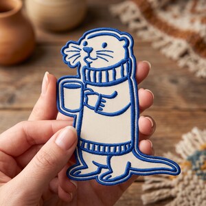 May include: An embroidered patch featuring a cream-colored otter wearing a blue sweater and holding a mug. The design is outlined in bold blue thread, showcasing the otter's whiskers, facial features, and the ribbed texture of the sweater collar and hem.