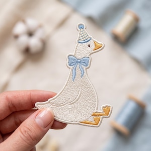 May include: An embroidered patch featuring a white goose wearing a light blue and white striped party hat and a light blue bow tie around its neck. The goose has a yellow beak and yellow webbed feet, rendered in detailed thread work.