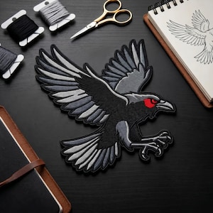 May include: Embroidered patch featuring a black raven in flight with gray and white wing details and red eyes. The patch is on a black surface, with spools of thread, gold scissors, and a sketchbook visible.