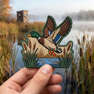 May include: An embroidered patch featuring a mallard duck in flight over a blue pond with green cattails. The duck has a green head, brown chest, tan body, and brown wings with a blue speculum. The patch has a dark green border and orange feet.