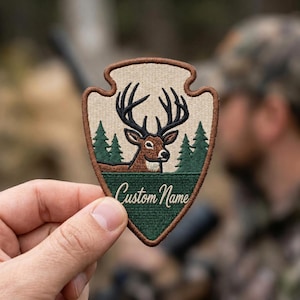 May include: An arrowhead-shaped embroidered patch featuring a brown deer with large antlers, dark green pine trees, and a cream-colored background. The lower section is dark green with the white cursive text Custom Name and a brown border.