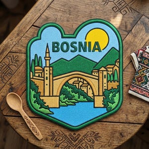 May include: A Bosnia patch with a green border, featuring a bridge, mountains, and a yellow sun against a blue sky. The word "BOSNIA" is across the top. A wooden spoon and a patterned textile are also visible.