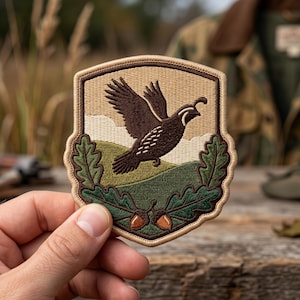 May include: A shield-shaped embroidered patch featuring a dark brown quail in flight against a tan sky and green rolling hills. The bottom of the patch is decorated with two green oak leaves and two brown acorns, all outlined in a dark brown border.