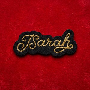 May include: A black felt patch with the name "Sarah" embroidered in gold thread. The name is in a cursive script and is outlined by a gold border. The patch is set against a red background.
