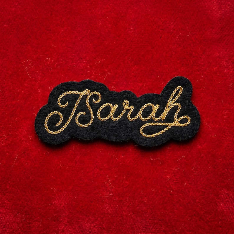 May include: A black felt patch with the name "Sarah" embroidered in gold thread. The name is in a cursive script and is outlined by a gold border. The patch is set against a red background.
