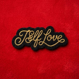 May include: A black embroidered patch with the words "Self Love" in gold script. The patch has a cloud-like shape and is set against a vibrant red background. The gold thread creates a textured, elegant look.