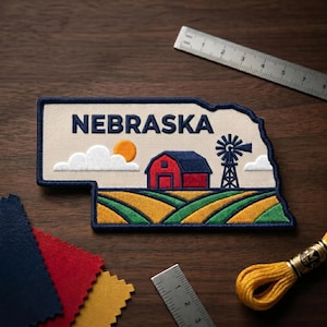 May include: Embroidered patch in the shape of Nebraska, featuring a red barn, windmill, sun, and fields. The word "NEBRASKA" is embroidered in navy blue. The patch is bordered in navy blue and set against a tan background.