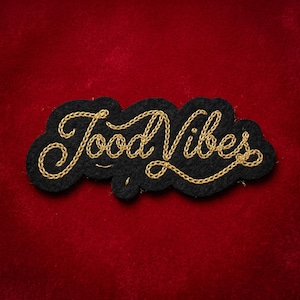May include: A black felt patch with the words "Good Vibes" embroidered in gold thread. The patch has a cloud-like shape and is set against a red velvet background.