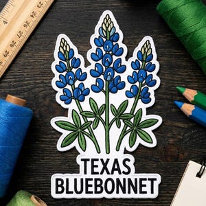 May include: An embroidered patch featuring three bluebonnet flowers with blue petals, green stems, and green leaves. Below the flowers, a white rectangular banner contains the black text TEXAS BLUEBONNET. A wooden ruler is visible on the left.