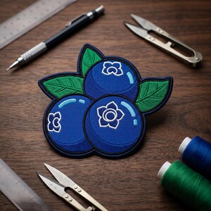May include: Embroidered patch featuring three blue blueberries with white and blue accents and green leaves. The patch is on a wooden surface with a pencil, scissors, and spools of thread.