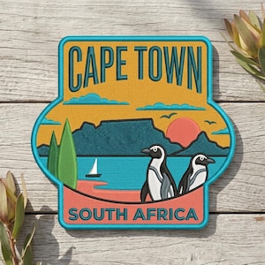 May include: Embroidered patch featuring a Cape Town, South Africa design. The patch has a teal border and includes the text "CAPE TOWN" and "SOUTH AFRICA". The design depicts a sunset, penguins, and a sailboat.