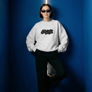 May include: A white sweatshirt with the text "Stay Wild" in a black, stylized font. The sweatshirt is paired with black pants. The person in the image is wearing sunglasses and leaning against a blue wall.