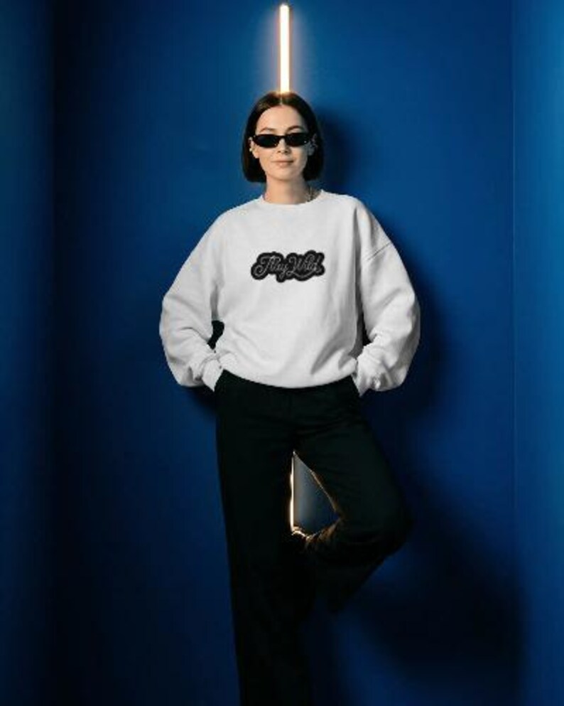 May include: A white sweatshirt with the text "Stay Wild" in a black, stylized font. The sweatshirt is paired with black pants. The person in the image is wearing sunglasses and leaning against a blue wall.