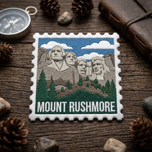 May include: Embroidered patch featuring Mount Rushmore with a blue sky background and the text "MOUNT RUSHMORE". The patch has a white border and is surrounded by pine cones, a compass, and a leather-bound journal.