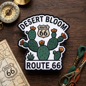 May include: An embroidered patch featuring a green cactus with orange flowers on a white background. The text reads DESERT BLOOM and ROUTE 66, with a Route 66 shield icon in the center. The patch has a black border and is shaped with a curved top.