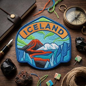 May include: An embroidered patch featuring a landscape of Iceland with a central orange lava flow, a snow-capped mountain, and green aurora borealis against a light blue sky. The word ICELAND is stitched in blue letters on an orange banner.