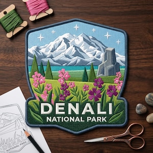 May include: Embroidered patch featuring a mountain landscape with the text "DENALI NATIONAL PARK." The patch includes a blue sky with stars, a mountain range, green trees, and pink and purple flowers. The patch is surrounded by a dark green border.